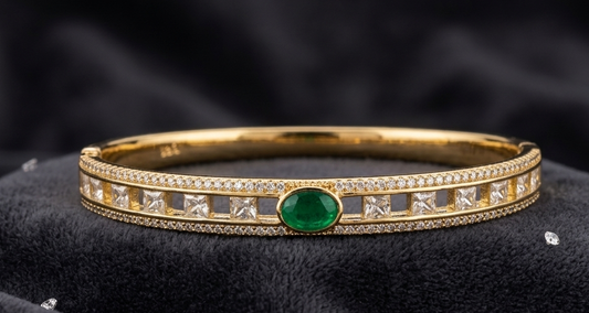 A gold bangle bracelet featuring a central oval green emerald and square-cut clear stones, displayed on a black velvet fabric background.