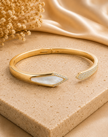 Gold open cuff bangle bracelet with a mother-of-pearl arrowhead inlay and pavé-set Cubic Zircon Triple "A" Quality on one end, displayed on a beige stone block with gold-toned fabric and dried flowers in the background.