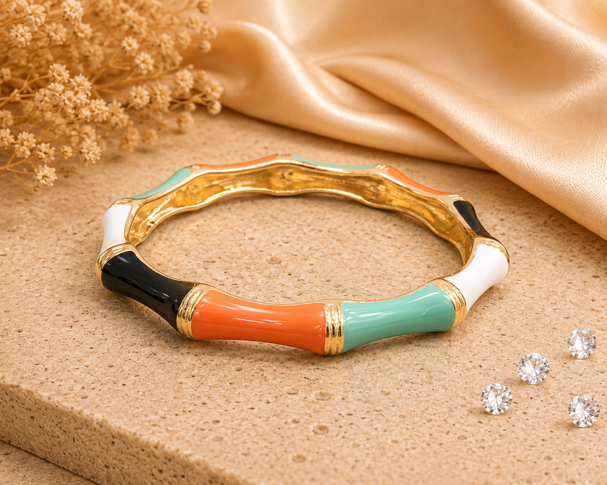 A colorful enamel bangle bracelet styled on a beige stone surface with draped champagne silk fabric and dried flowers. The bracelet features segments of orange, mint green, white, and black with gold trim.