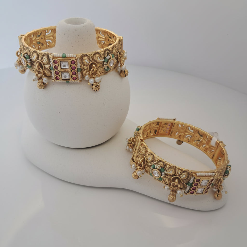 Two ornate gold bracelets with gemstones on a white stand against a light gray background.