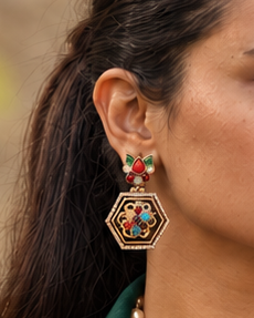 A woman wearing a dark green double-breasted blazer, a long cream pearl necklace with a geometric pendant, and matching geometric earrings, posed against a blurred stone wall background.