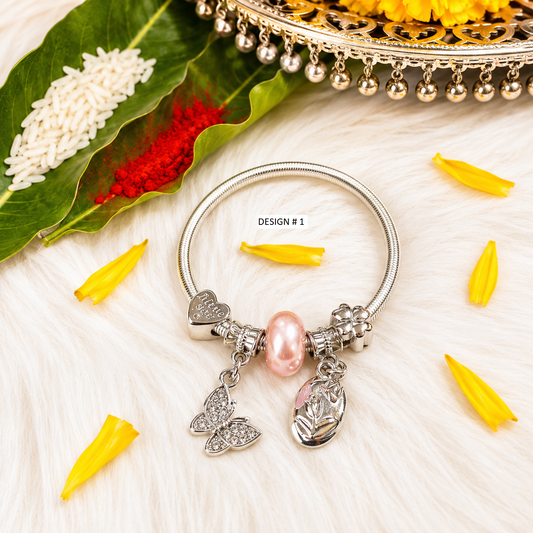 A silver-toned bangle bracelet with dangling charms, including a pink pearl, a butterfly, and a lotus flower, displayed on a white faux fur surface with decorative yellow petals, a green leaf, and a gold tray in the background.