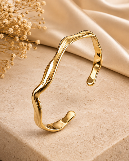 A polished gold open cuff bracelet with an organic, wavy fluid design, displayed on a beige stone block with a draped satin fabric and dried baby's breath flowers in the background.