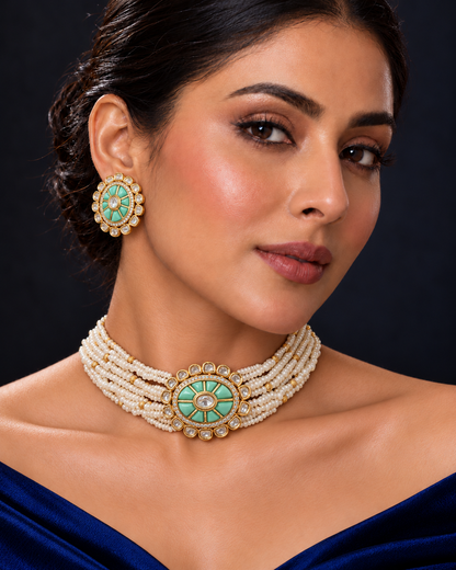 A close-up of a woman wearing a multi-strand pearl choker necklace with a turquoise and gold circular centerpiece, matching round turquoise earrings, and a dark blue off-the-shoulder dress against a dark background.