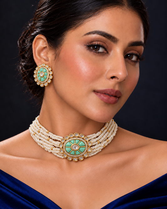 A close-up of a woman wearing a multi-strand pearl choker necklace with a turquoise and gold circular centerpiece, matching round turquoise earrings, and a dark blue off-the-shoulder dress against a dark background.