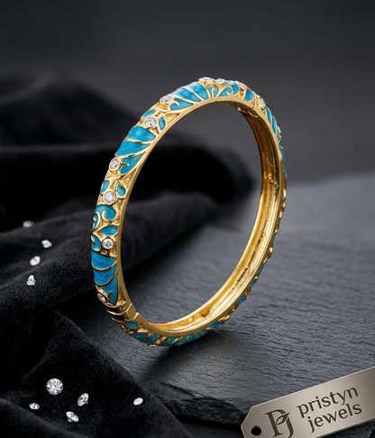 A gold-plated bangle bracelet with intricate turquoise blue enamel leaf patterns and small clear rhinestone accents, displayed on a dark textured fabric background with a branded tag.