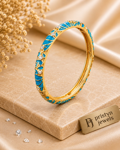 A gold bangle bracelet with bright blue enamel inlay and small Cubic Zircon Triple "A" Quality, displayed on a beige textured block with dried flowers and satin fabric in the background. A small card reading 'pristyn jewels' is visible at the lower right.