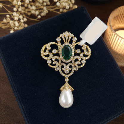 An ornate gold pendant brooch featuring a green oval stone, clear rhinestones, and a dangling white pearl, displayed on a dark velvet box with a handwritten price tag.
