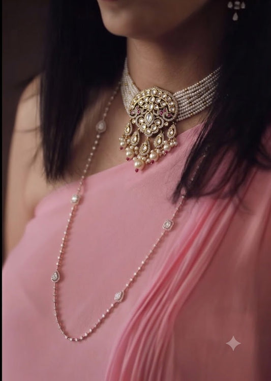 A close-up of a woman wearing traditional Indian jewelry, featuring a gold and pearl choker necklace with a large central pendant and a matching long pearl strand draped over a pink draped dress.
