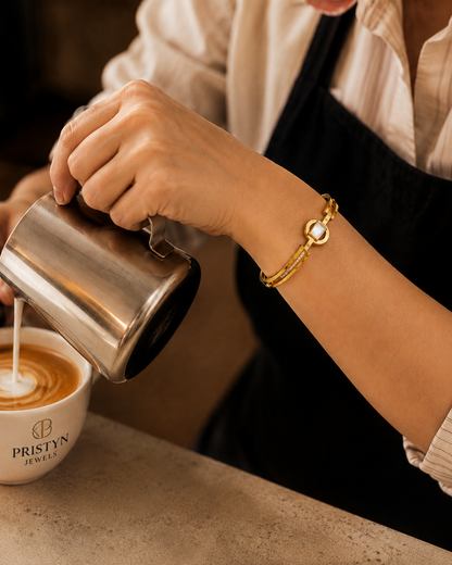 A close-up lifestyle shot of a barista pouring milk into a cup of latte art. The barista is wearing a gold bangle bracelet with a white pearl and gold chain, along with a black apron over a cream-colored shirt.