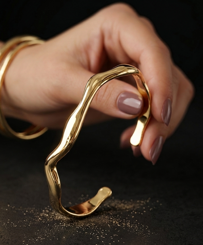 A hand with mauve nail polish holds a polished gold cuff bracelet with a wavy, organic curve. Another set of gold bangles is visible on the wrist, set against a dark textured background with scattered gold glitter.