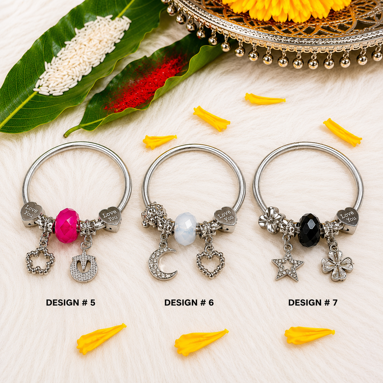Three silver-toned ring charms labeled DESIGN #5, DESIGN #6, and DESIGN #7 are arranged on a white textured surface with decorative green leaves, yellow flower petals, and a gold tray in the background. The rings feature different dangling charms: a pink bead and moon charm in Design 5, a white pearl and crescent moon charm in Design 6, and a black bead and star/floral charms in Design 7.