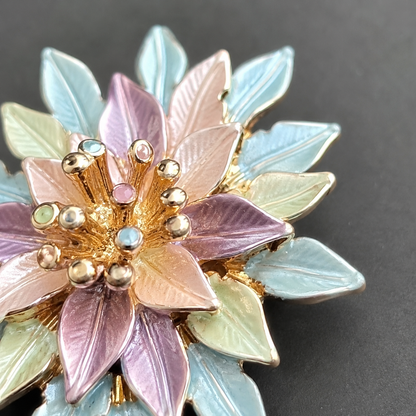A close-up of a floral enamel brooch with pastel purple, light blue, and pale green petals, accented by gold-tone metal and small iridescent rhinestones, set against a dark gray background.