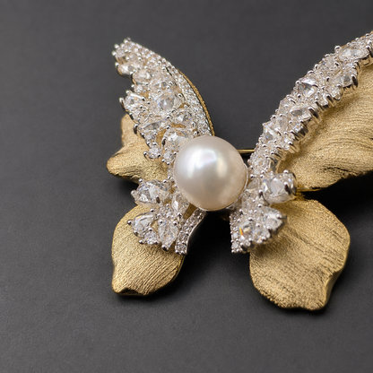 A butterfly-shaped brooch featuring textured gold-tone wings, clear rhinestone-encrusted sections, and a central white pearl, photographed on a dark gray background.