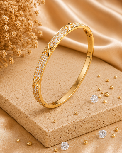 A gold bangle bracelet with pavé-set Cubic Zircon Triple "A" Quality and geometric diamond-shaped accents, displayed on a textured beige stone block with gold fabric and dried floral props.