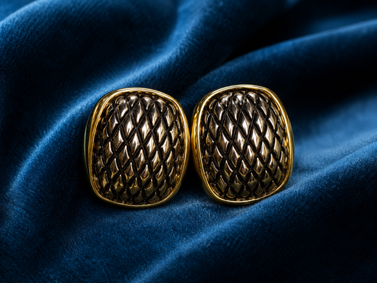 A pair of gold-toned square cufflinks with an intricate raised geometric lattice pattern, displayed on a draped dark blue velvet fabric.