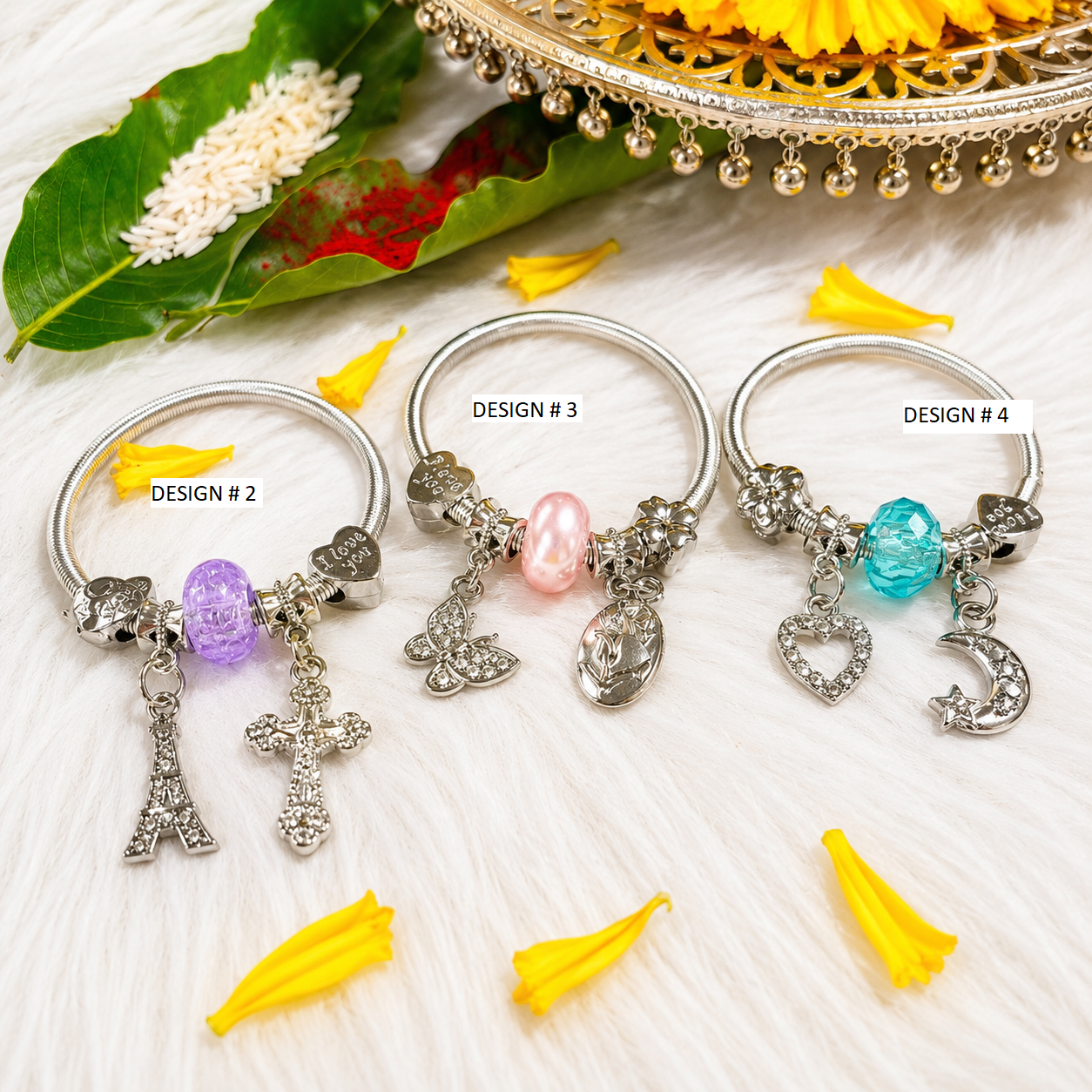 Four silver-toned bangles labeled DESIGN #2, DESIGN #3, DESIGN #4, displayed on a white faux fur surface with decorative yellow flower petals, a green leaf, and a gold-toned tray in the background. The bangles feature different charm combinations including crystals, a cross, a heart, a crescent moon, a butterfly, and colored beads in purple, pink, and blue.