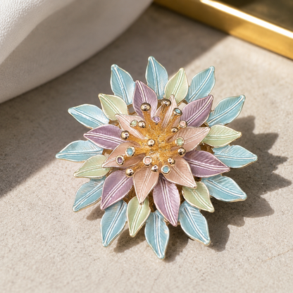 A floral enamel brooch with layered pastel petals in lavender, mint green, and light blue, surrounding a gold-toned center with small clear rhinestones, photographed on a textured beige surface.