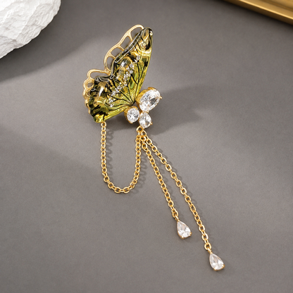 A gold butterfly-shaped brooch with green and gold enamel detailing, adorned with clear rhinestones and dangling gold chains with teardrop crystal charms, displayed on a dark gray surface.