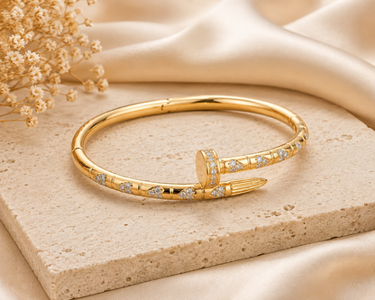 A gold hinged bangle bracelet with pavé-set Cubic Zircon Triple "A" Quality, styled on a beige stone slab with cream satin fabric and dried flowers in the background.