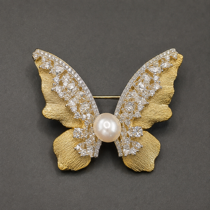 A butterfly-shaped brooch in textured yellow gold, embellished with pavé-set clear crystals along the wings and a central round pearl, photographed on a dark gray background.