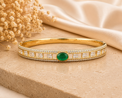 A gold bangle bracelet with a central oval green emerald stone and square-cut Cubic Zircon Triple "A" Quality along the band, styled on a beige stone surface with cream fabric and dried flowers.