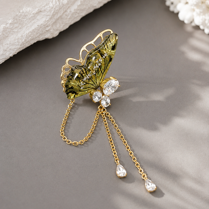 A gold butterfly-shaped hair claw pin with green enamel wings and clear rhinestones, featuring dangling gold chains with teardrop rhinestone pendants, displayed on a gray surface.