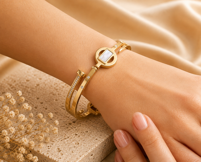 A close-up of a person's wrist wearing a gold-toned multi-band bracelet with a central oval link featuring a white inlay, accented by a slim bangle with small Cubic Zircon Triple "A" Quality, set against a warm beige satin and stone background.