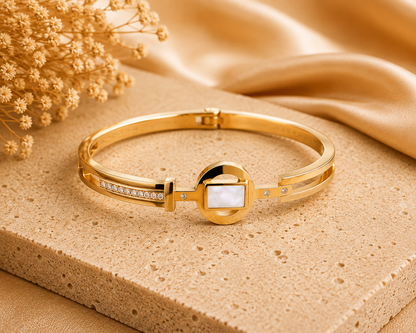 A gold-plated bangle bracelet with a central white inlay and small Cubic Zircon Triple "A" Quality, displayed on a textured beige stone slab with gold satin fabric and dried flowers in the background.