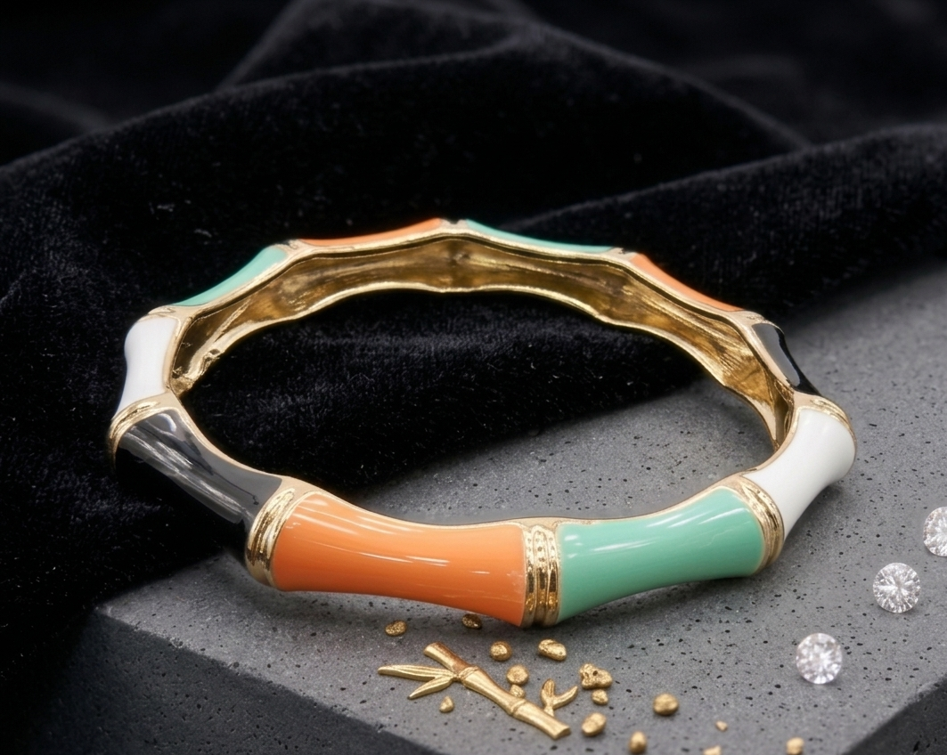 A gold-plated bangle bracelet with a bamboo-inspired segmented design, featuring glossy enamel sections in orange, mint green, white, and black, displayed on a dark textured surface with scattered gold beads and diamonds.