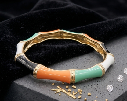 A gold-plated bangle bracelet with a bamboo-inspired segmented design, featuring glossy enamel sections in orange, mint green, white, and black, displayed on a dark textured surface with scattered gold beads and diamonds.
