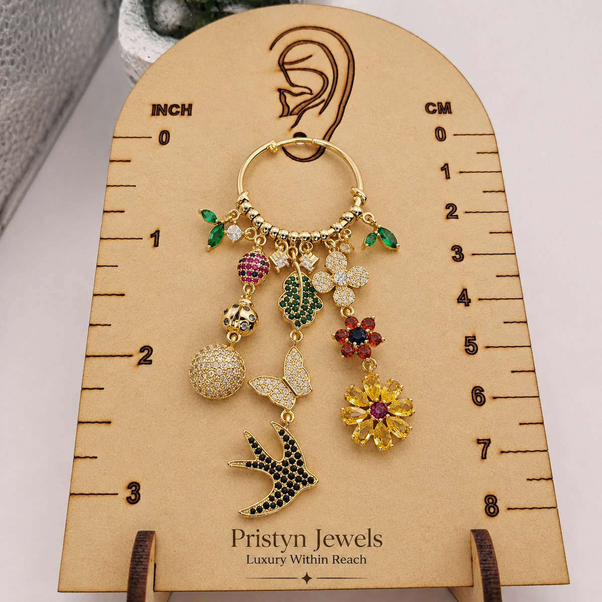A gold-toned hoop earring with dangling charm elements including green leaves, pink and red flowers, a white butterfly, a yellow flower, a bird, and a round pavé bead, displayed on a beige measurement card labeled for earring size. The card shows the brand name 'Pristyn Jewels'.