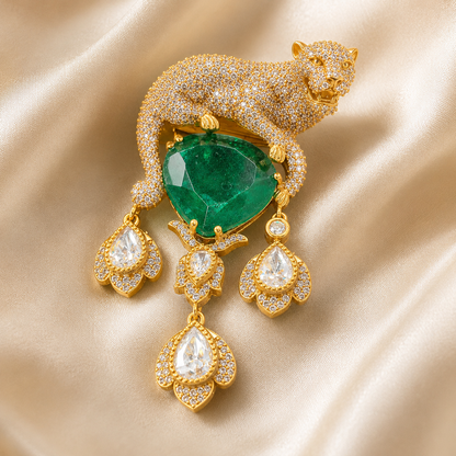 A luxury gold-toned jewelry piece featuring a panther-shaped brooch adorned with pavé diamonds and a large heart-shaped green emerald, with three matching diamond-detailed teardrop dangles, displayed on a beige satin fabric background.
