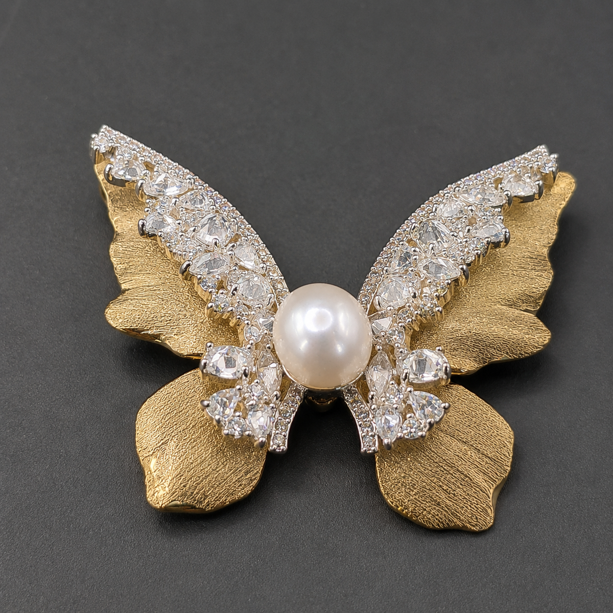 A butterfly-shaped brooch featuring textured gold-tone wings, a central white pearl, and clear rhinestone-encrusted wings, displayed on a dark gray background.