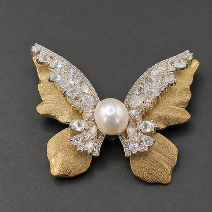 A butterfly-shaped brooch featuring textured gold-tone wings, a central white pearl, and clear rhinestone-encrusted wings, displayed on a dark gray background.