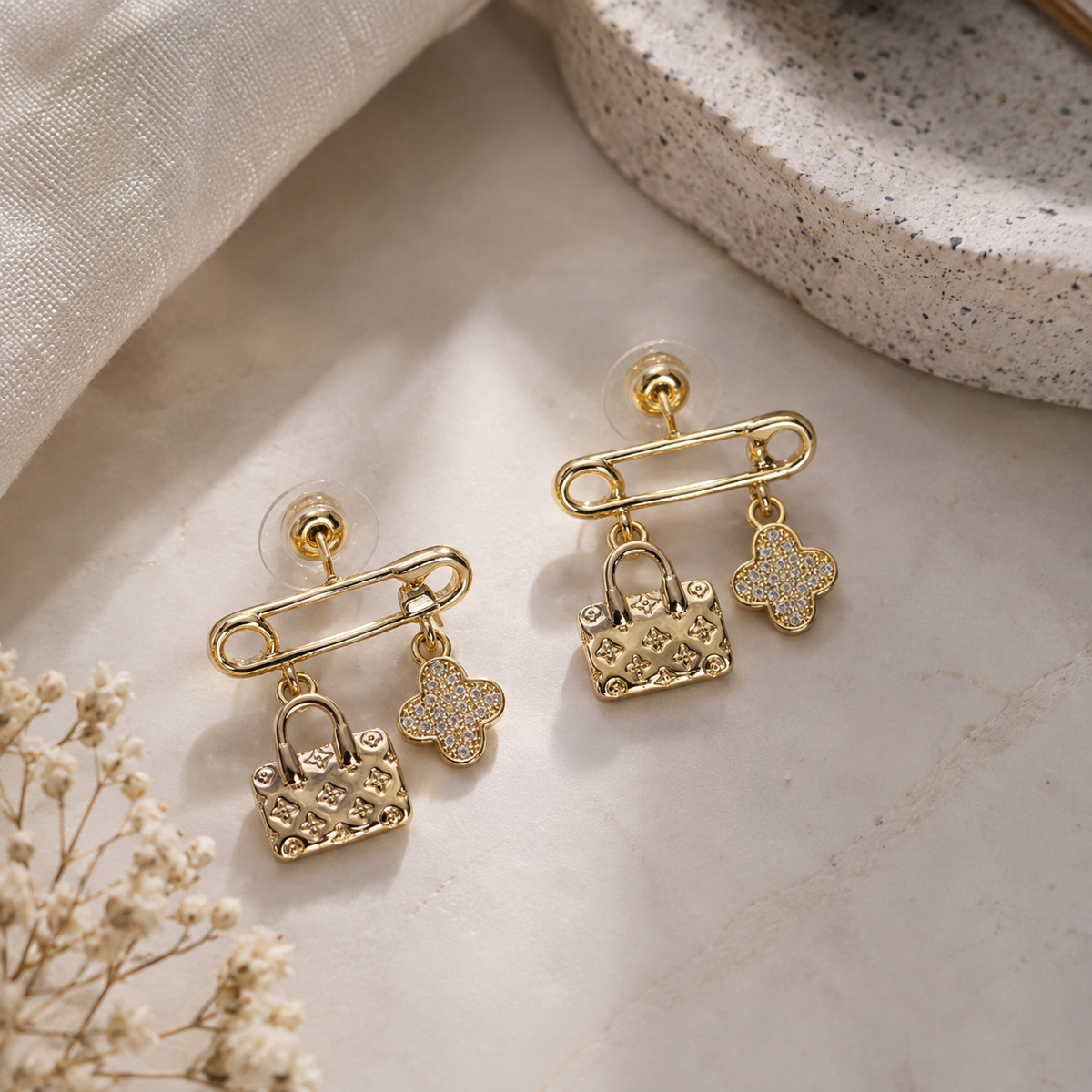 A pair of gold-toned safety-pin stud earrings with dangling textured lock charms and small flower-shaped crystal charms, displayed on a light marble surface with neutral props.