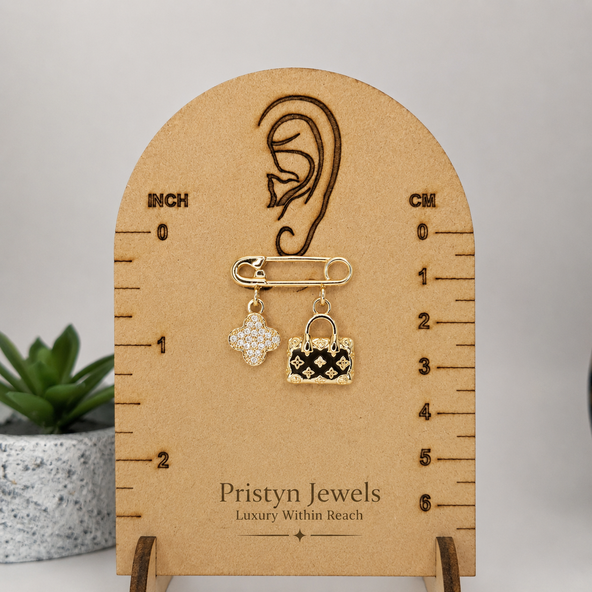 A gold-toned safety pin earring with two dangling charms, one rhinestone-encrusted and one black-and-gold patterned, displayed on a branded wooden ear-measuring stand. A small potted succulent sits at the left edge against a light gray background.