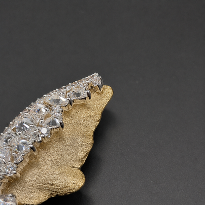 A close-up of a gold-toned leaf-shaped brooch or pin embellished with a curved row of clear gemstones in a silver-toned setting, photographed on a dark gray background.
