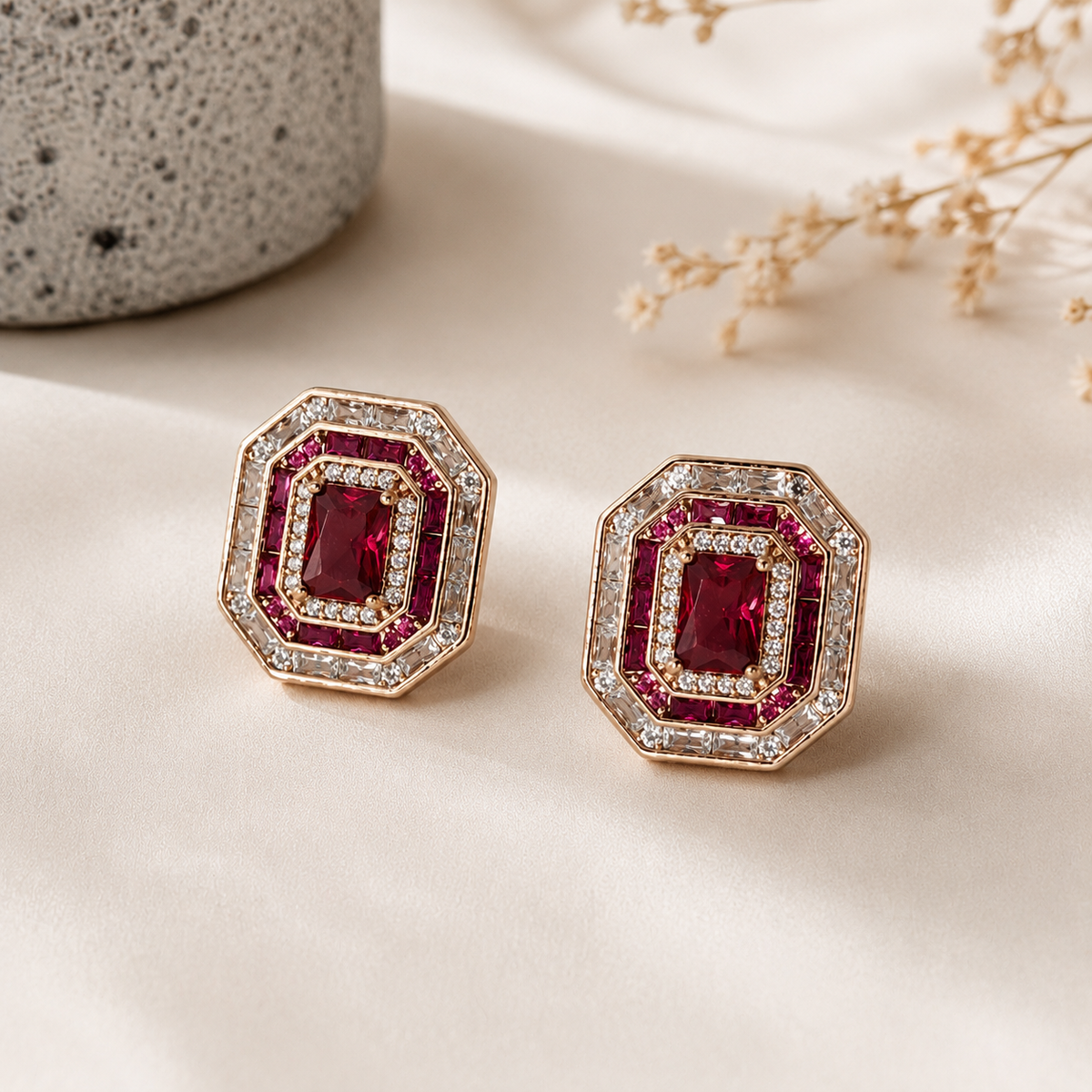 A pair of geometric stud earrings with rectangular red gemstones surrounded by Cubic Zircon Triple "A" Quality and pinkish-red accents, set in a gold-tone metal, displayed on a cream satin fabric with a textured ceramic object in the background.