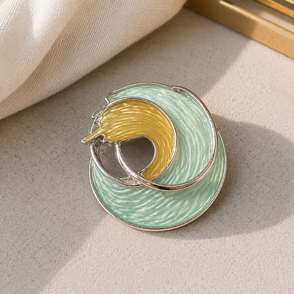 A close-up of a circular enamel brooch with a mint green outer ring, a pale yellow inner crescent, and silver-tone metal edging, resting on a light beige textured surface with a soft fabric prop in the corner.