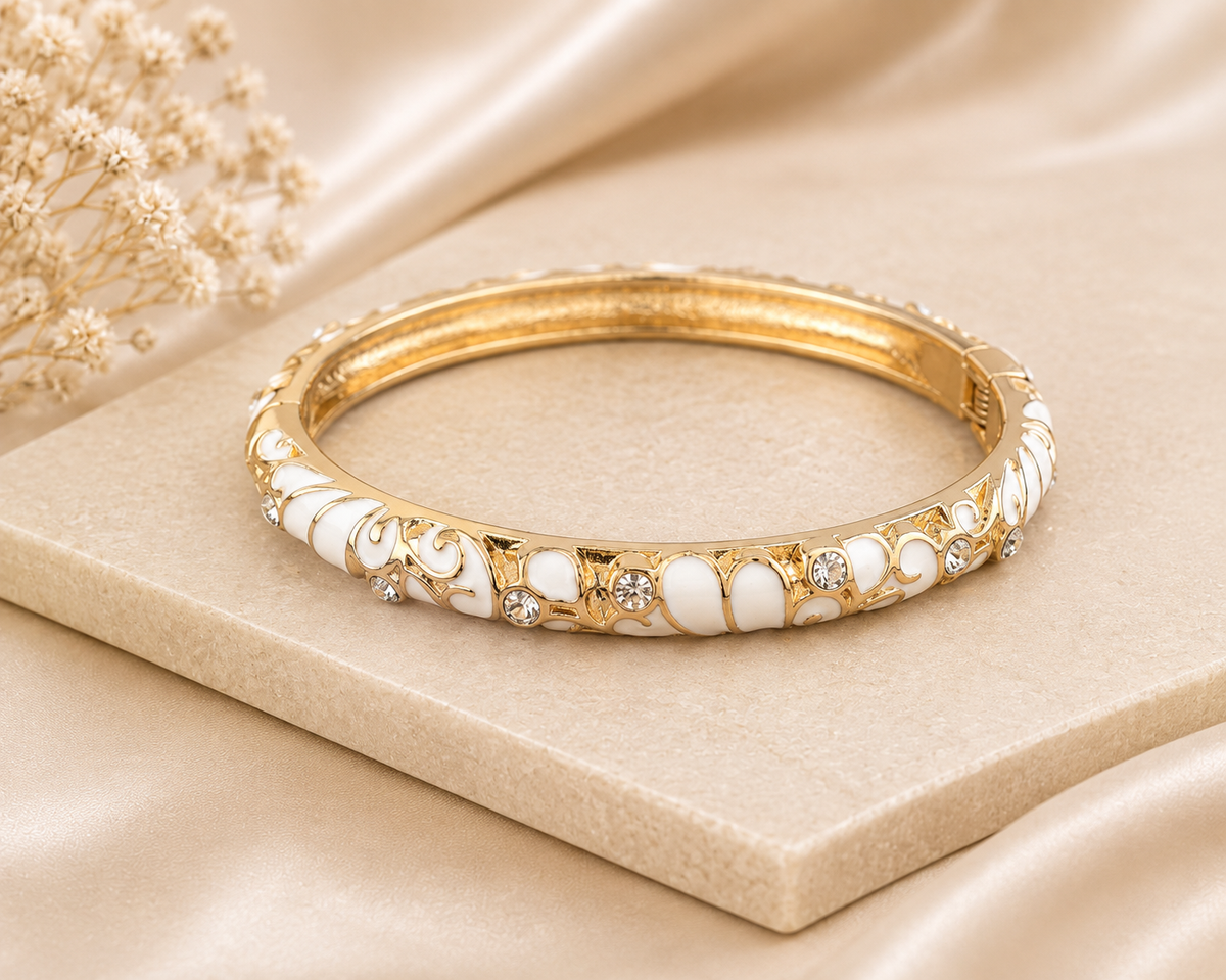 A gold-plated bangle bracelet with white enamel inlay and small clear crystal accents, displayed on a beige satin fabric background.